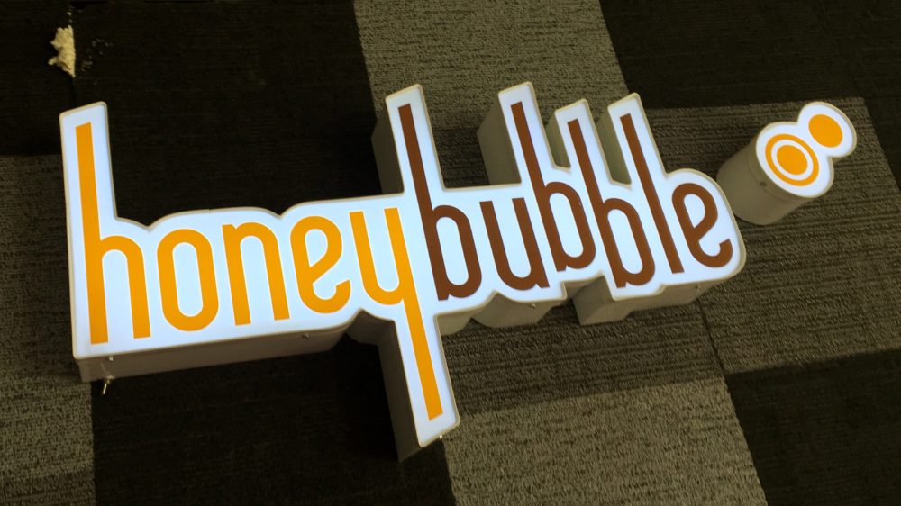 A neon logo sign for Honey Bubble Tea in the Virginia Highlands neighborhood of Atlanta Georgia. Yellow letters say honey and brown letters say bubble, on a white background, against a wall with brown squares