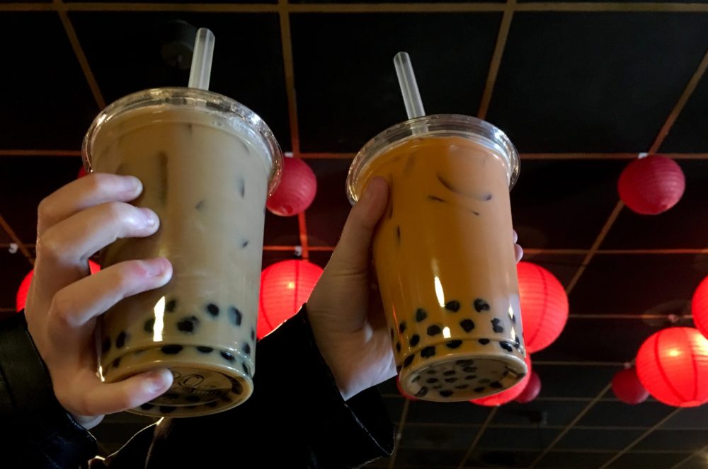 Two brown cups of bubble milk tea with brown tapioca balls in the bottom, being held by two hands in front of a black ceiling decorated with red asian lanterns