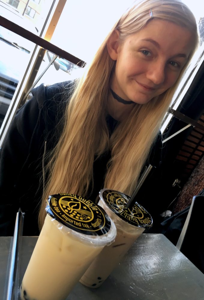 Trillian Celeste, a girl dressed in black with long blonde hair sitting behind two cups of light brown bubble tea with black tapioca boba balls in the bottom of each