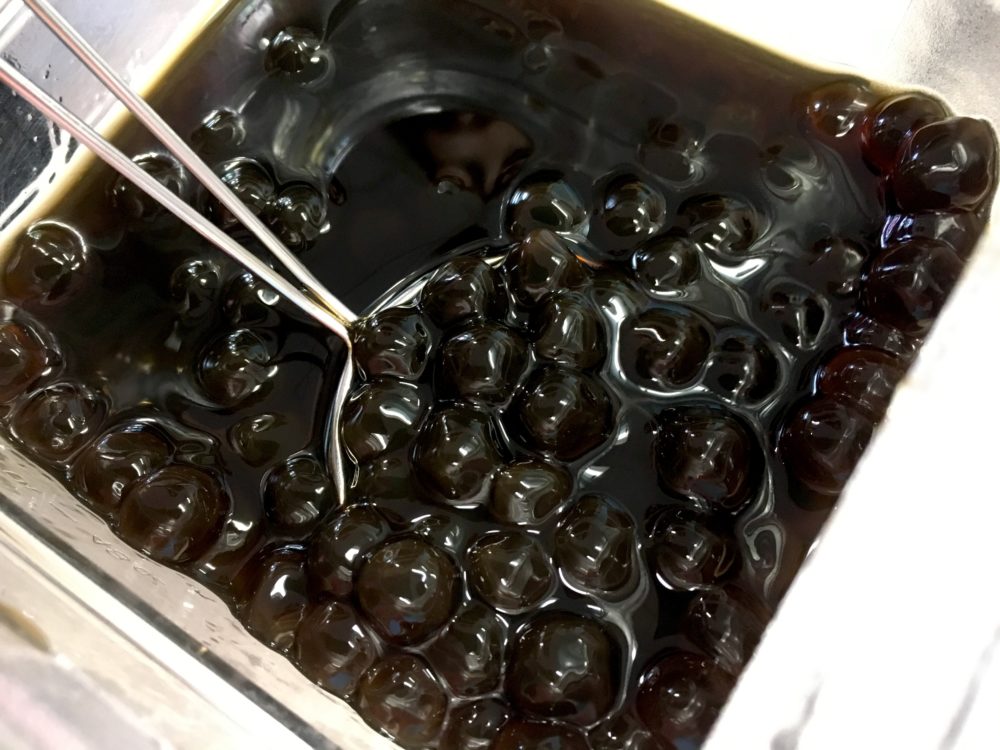 A clear acrylic serving container filled with shiny black tapioca pearls. This is a self-serve boba topping station at WIN Bubble Tea in Nashville, Tennessee.
