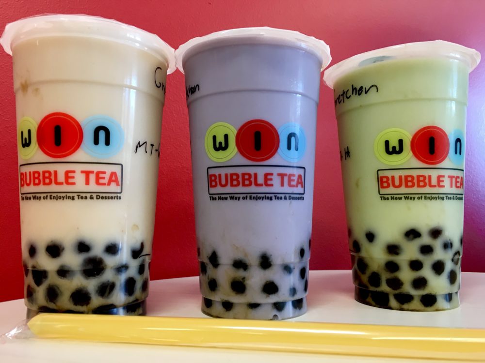 Three cups of bubble tea with tapioca boba balls. Flavors are cream colored wintermelon, purple taro, and green honeydew. Cups are against a pink wall with yellow straws in front.