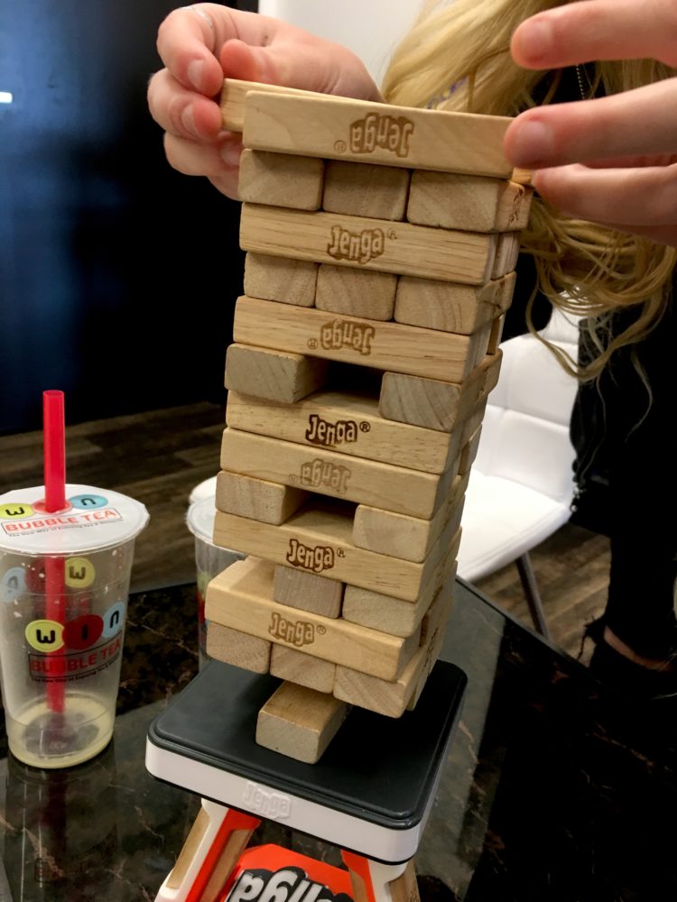 Trillian Celeste, a young girl with long blonde hair, uses her fingers to stack wooden rectangular JENGA game pieces in the game lounge of red Harrah's Casino poker playing cards and red, white, and blue round poker chips in the foreground. This is on the table in the game lounge of WIN Bubble Tea in Nashville, Tennessee. An empty bubble tea cup is in the background.
