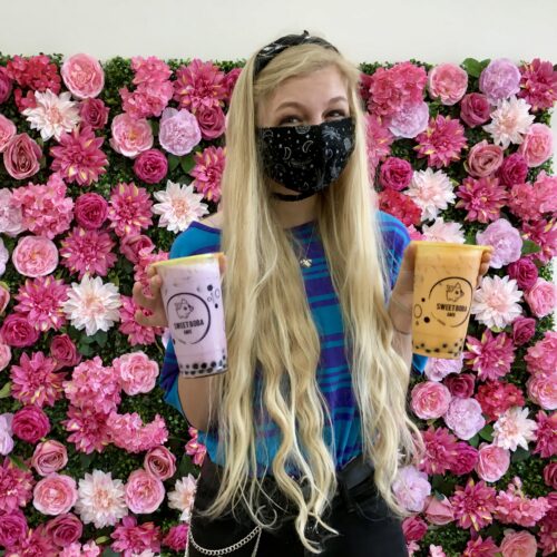 Young girl in a blue striped shirt with very long light blonde hair wearing a black mask and black headband, standing in front of a pink flower filled background, holding a cup of purple taro coconut bubble tea in one hand, and an orange thai coconut bubble tea in the other hand. Both have black tapioca boba balls in the bottom.
