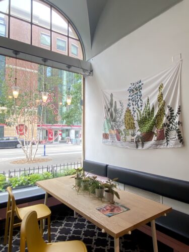 White Flag decorated with green plants, hanging above a light Brown Wooden Table and Seating Area in Coconut Whisk Bubble Tea Cafe in Minneapolis, Minnesota. All in front of a bright window in which you can see the street outside