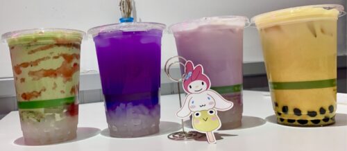 Four colorful cups of vegan bubble tea against a white wall. The fist cup is green and red strawberry matcha with clear coconut jelly, the next is a vivid purple butterfly pea tea with clear coconut jelly, the nest is a light purple taro tea with clear coconut jelly, and the last is a golden yellow turmeric tea with black tapioca pearls in the bottom. 