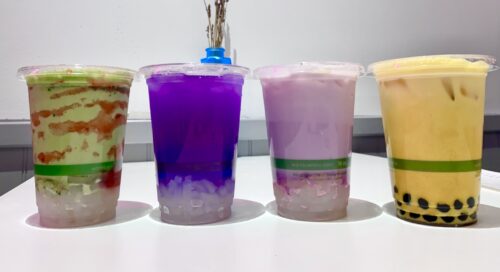 Four colorful cups of vegan bubble tea against a white wall. The fist cup is green and red strawberry matcha with clear coconut jelly, the next is a vivid purple butterfly pea tea with clear coconut jelly, the nest is a light purple taro tea with clear coconut jelly, and the last is a golden yellow turmeric tea with black tapioca pearls in the bottom. 