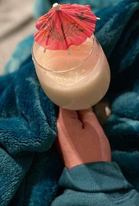 A Homemade Pina Colada in decorative glass to celebrate the New Year. This white drink is garnished with a red drink umbrella, on the backdrop of a fuzzy teal green blanket.